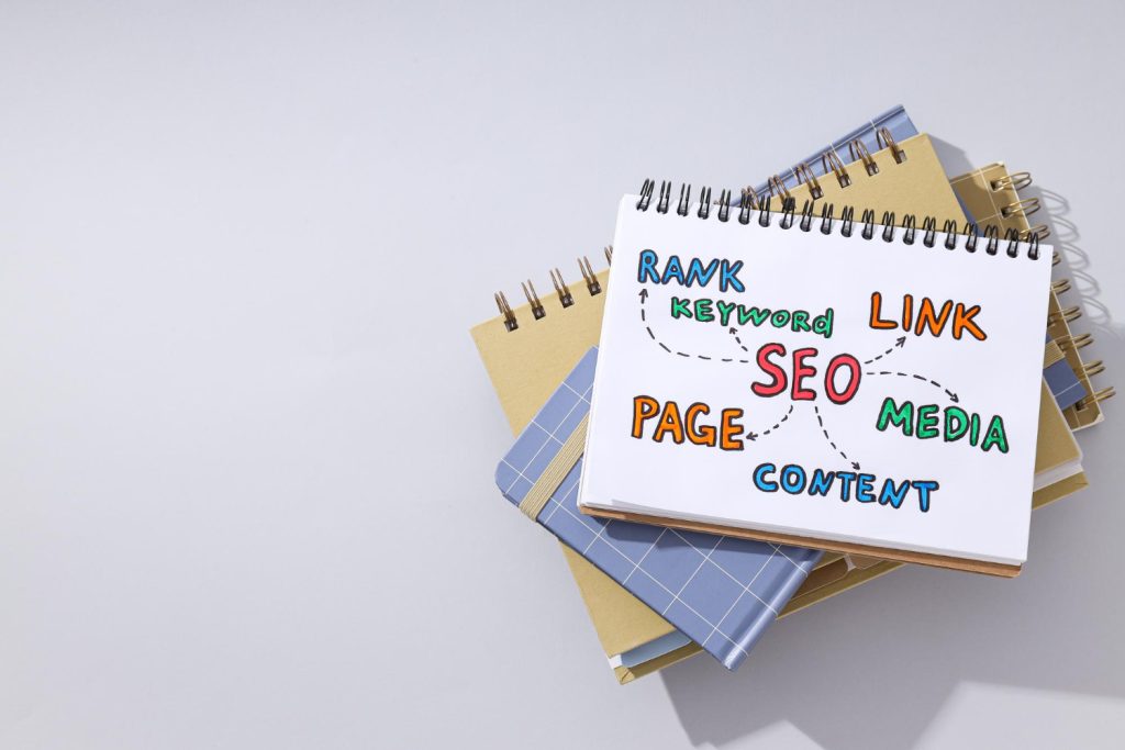 Un teanc de caiete cu o pagină de titlu pe care sunt scrise cu culori vii noțiuni de SEO precum „Rank”, „Link” și „Content”, legate între ele prin săgeți