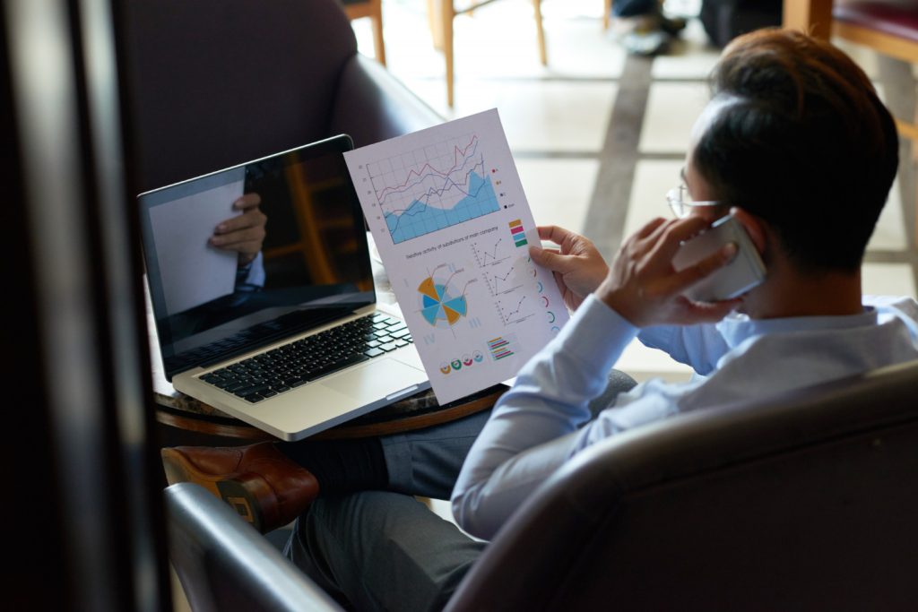 Vedere peste umăr a unui bărbat care efectuează multitasking și revizuiește documente financiare și are o conversație telefonică